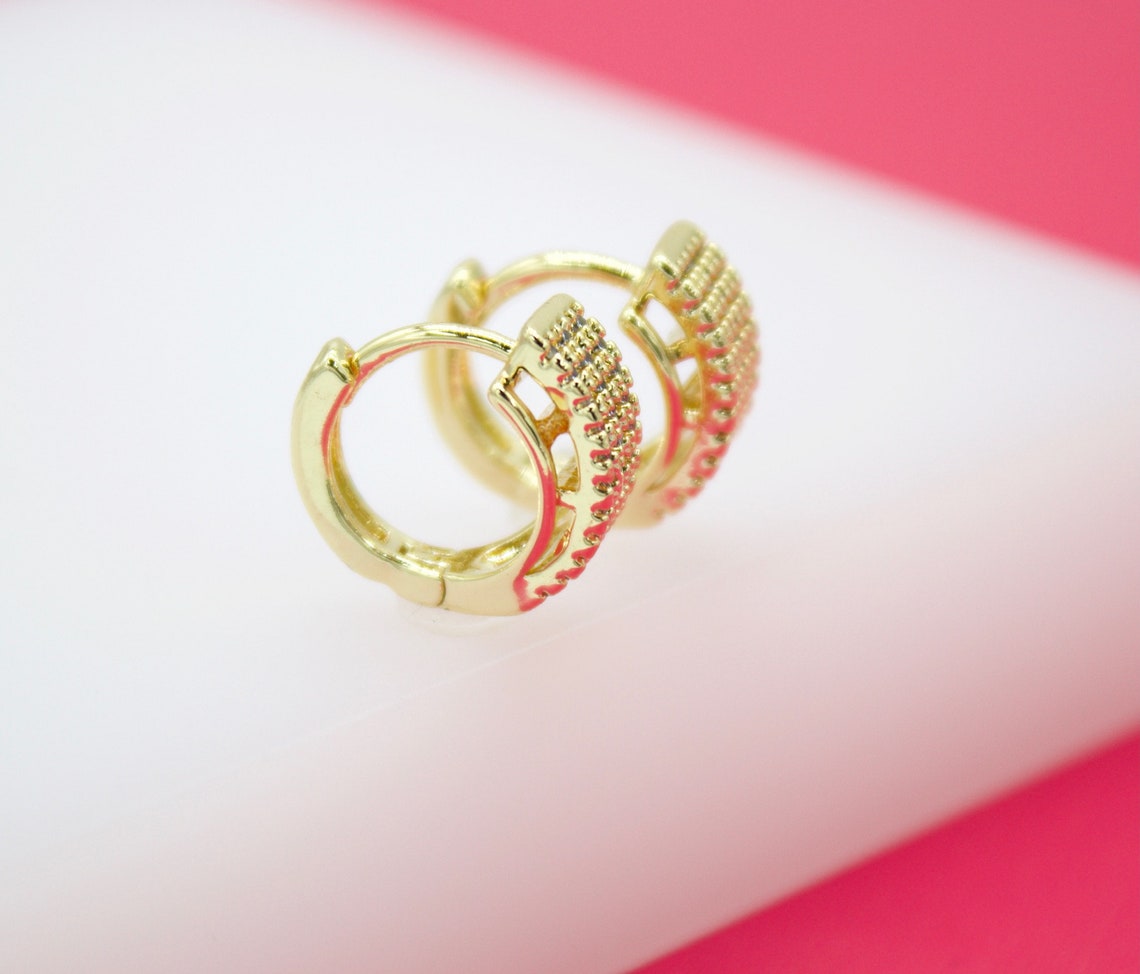 A pair of gold huggie hoop earrings with textured, studded details rests on a white and pink surface.