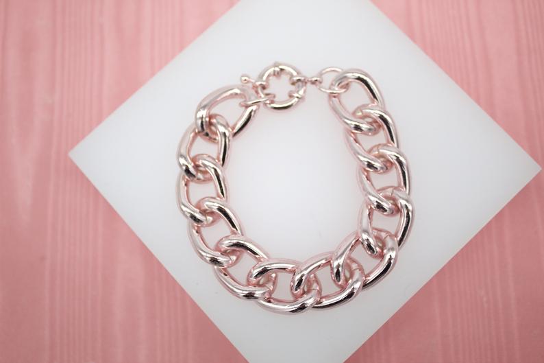 A chunky rose gold link bracelet rests on a white display card against a pink wooden backdrop.