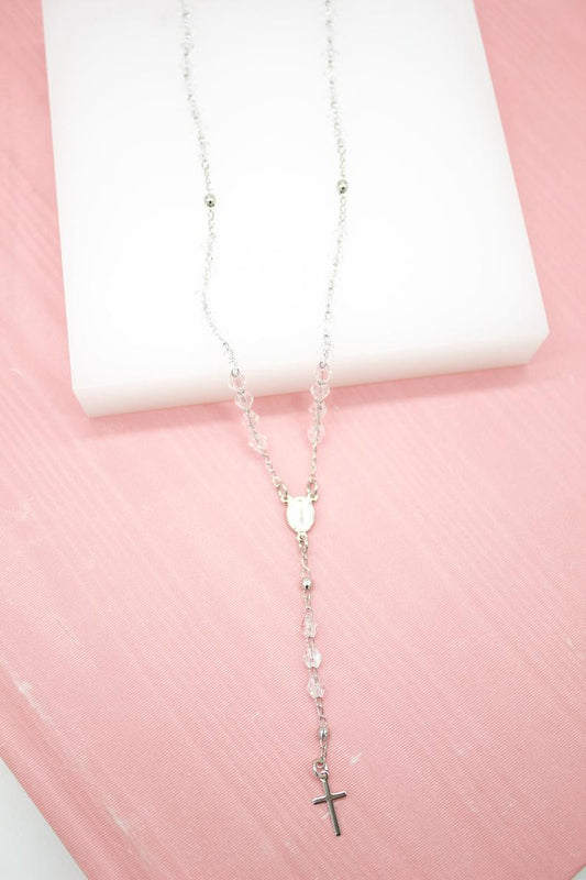 A delicate silver rosary necklace with crystal beads and a crucifix rests on a white display block against a soft pink background.