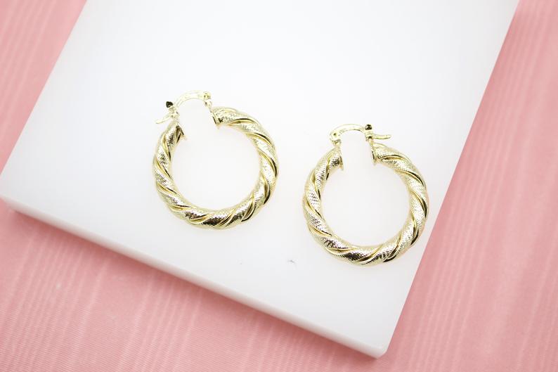 A pair of textured gold twist hoop earrings rests on a white display surface against a pink background.