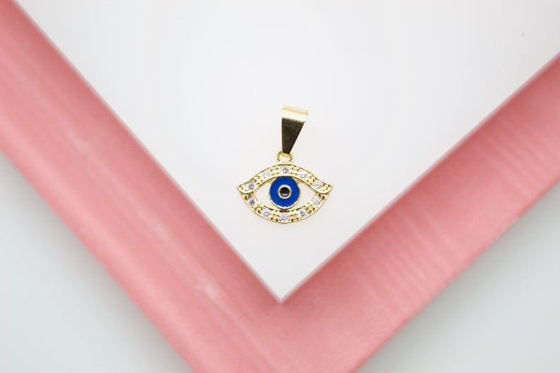 A gold evil eye pendant set with cubic zirconia stones and a bright blue enamel center rests on a white and pink surface.