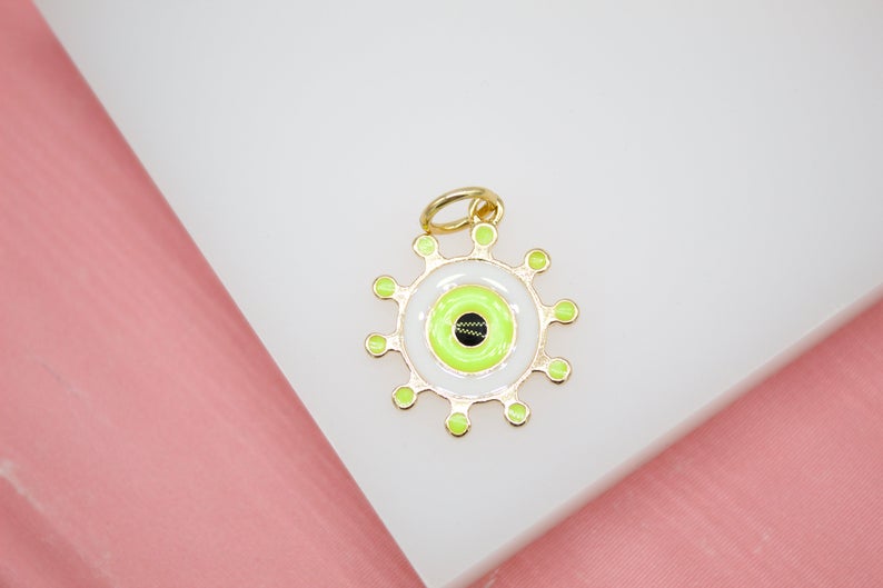 A gold evil eye charm featuring vibrant neon green and white enamel details rests on a white surface against a pink background.