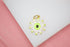 A gold evil eye charm featuring vibrant neon green and white enamel details rests on a white surface against a pink background.