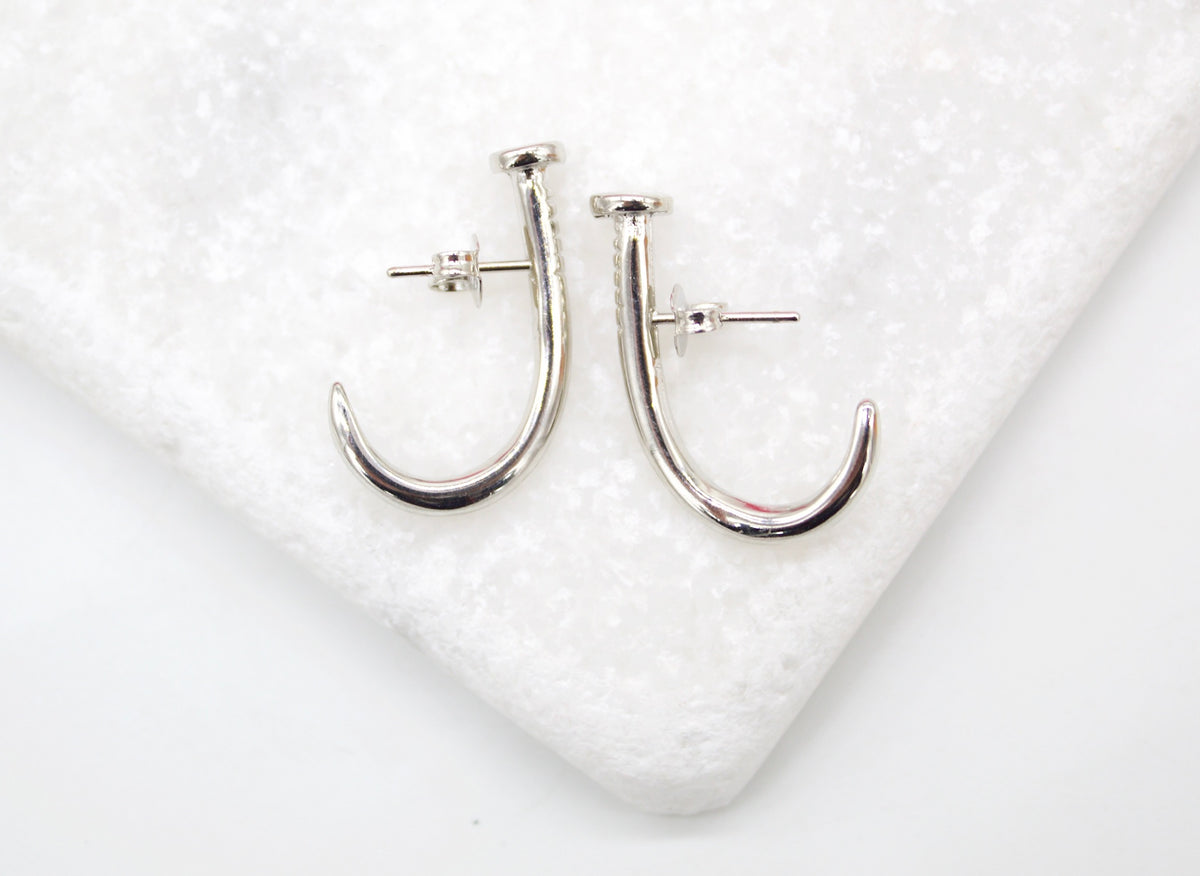 A pair of silver nail-shaped hoop earrings rests on a textured white surface.