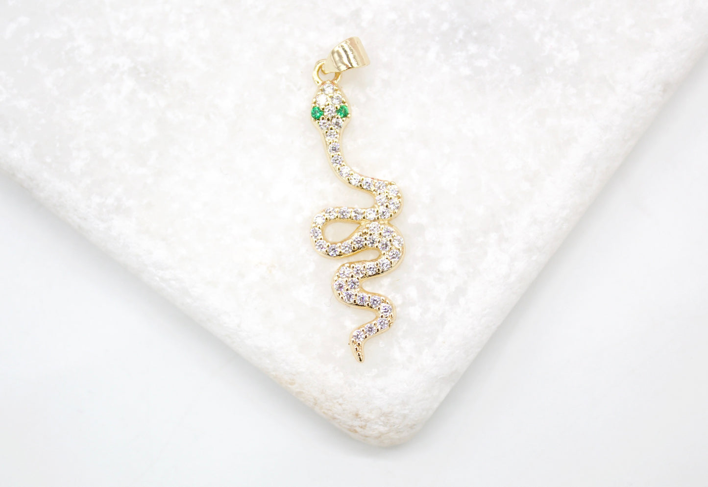 A sparkling gold snake pendant with diamond pave and green emerald eyes rests on a white marble surface.