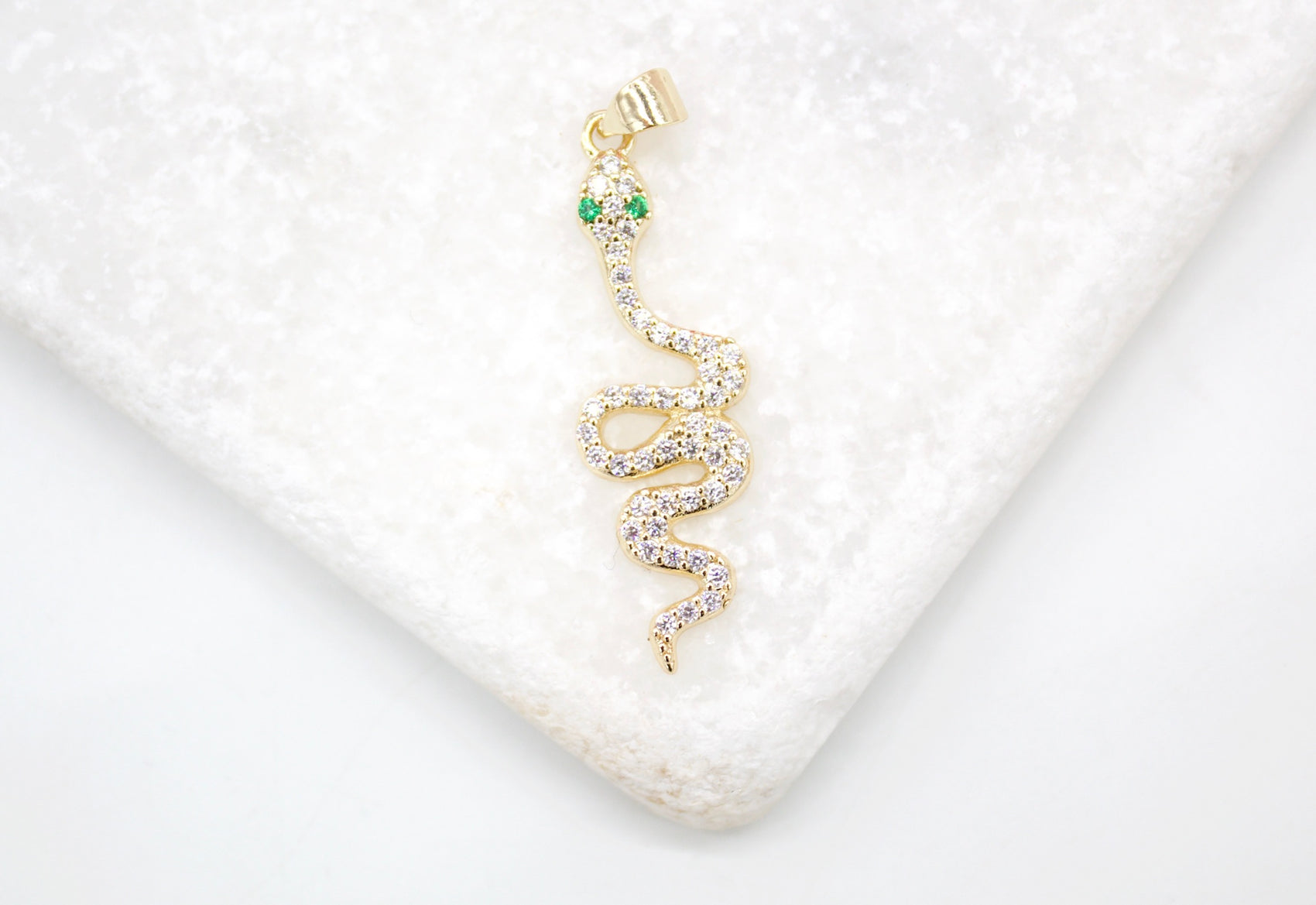 A sparkling gold snake pendant with diamond pave and green emerald eyes rests on a white marble surface.