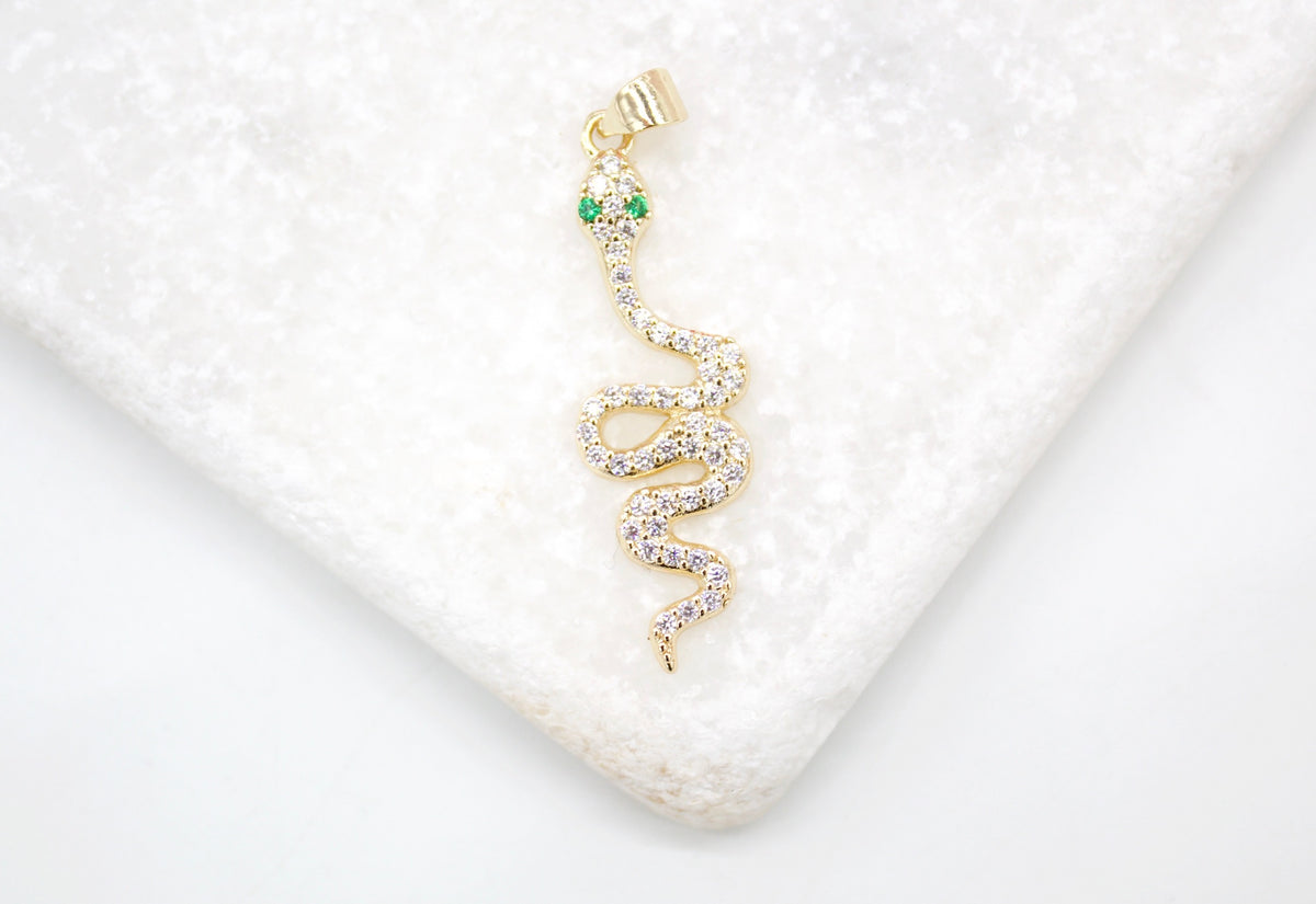 A sparkling gold snake pendant with diamond pave and green emerald eyes rests on a white marble surface.