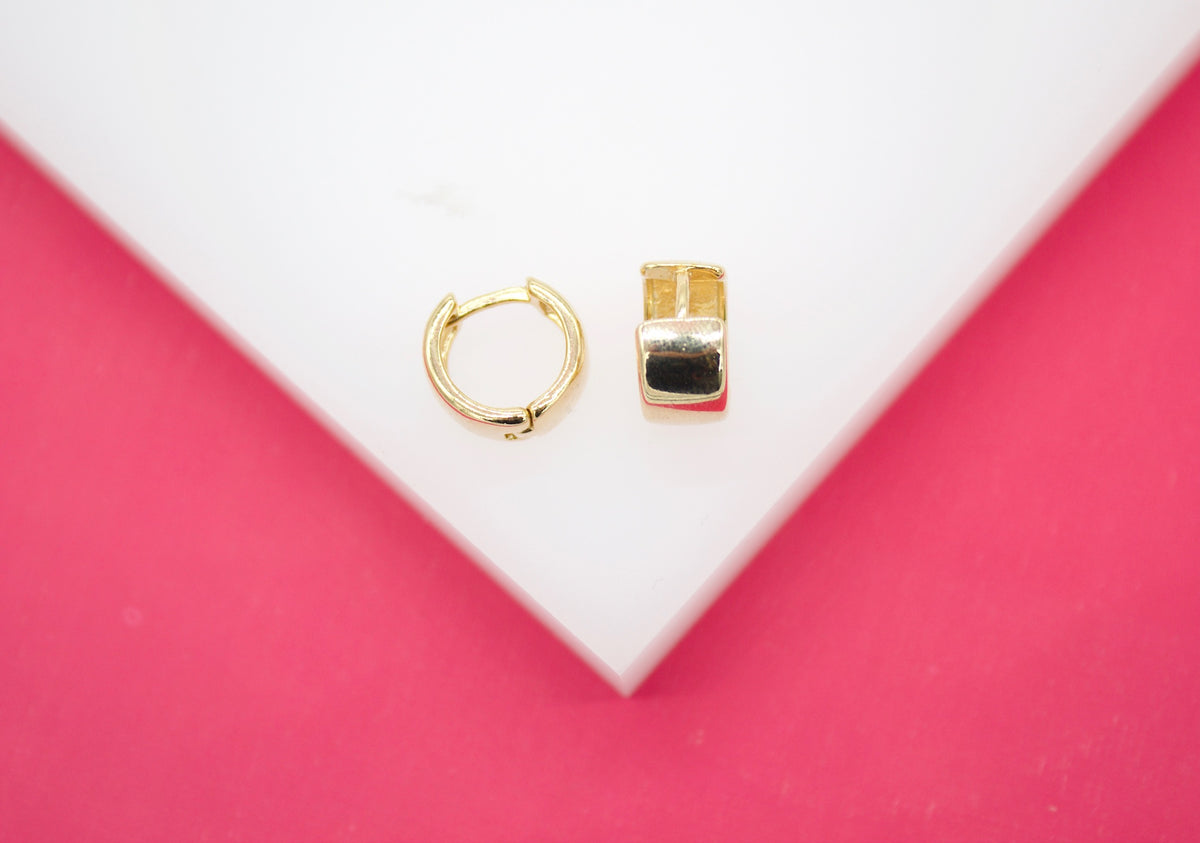 A pair of polished gold huggie hoop earrings rests elegantly on a white geometric display against a vibrant pink background.