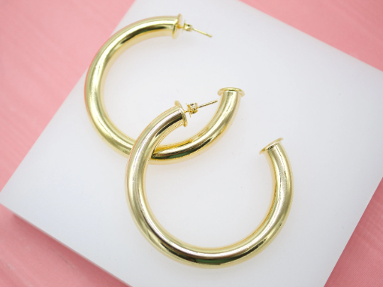 A pair of thick gold hoop earrings rests on a white display block against a pink background.