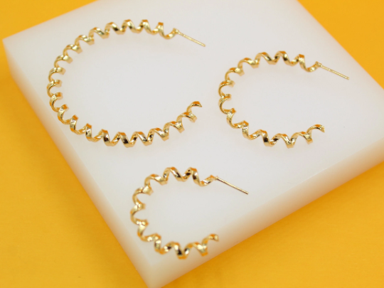 Three spiral gold hoop earrings of varying sizes rest on a white display stand against a bright yellow background.