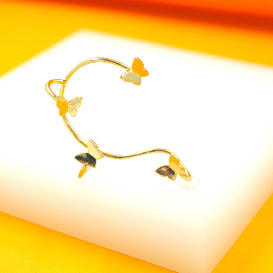 A delicate gold ear cuff featuring multiple colorful butterfly charms rests on a white display stand.