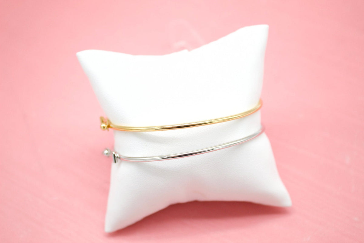 A gold bangle and a silver bangle rest elegantly on a white display pillow against a soft pink background.