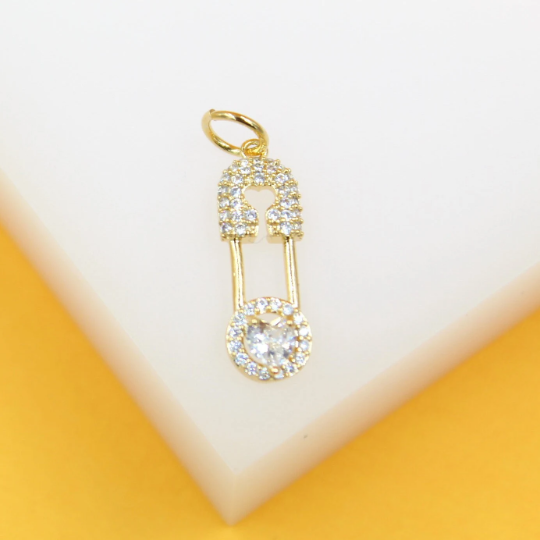 A gold safety pin pendant encrusted with sparkling cubic zirconia stones rests against a white and yellow background.