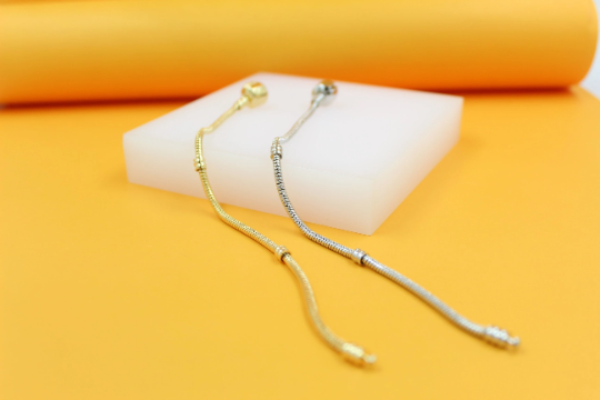 A gold and a silver snake chain bracelet rest diagonally on a white block against a bright yellow background.