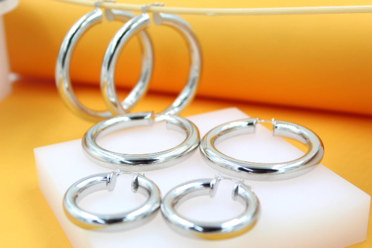 An assortment of thick silver hoop earrings in varying sizes sits on a white display block against an orange backdrop.