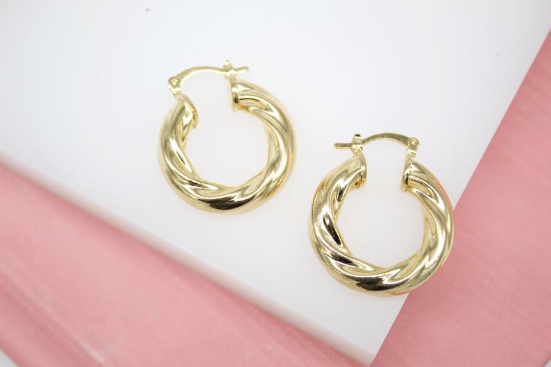 A pair of polished gold twist hoop earrings rest on a white surface next to a pink background.