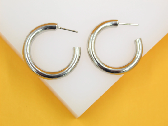 A pair of thick silver hoop earrings rests on a white display against a vibrant yellow background.