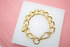 A chunky gold chain bracelet rests on a white display pad against a pink background.