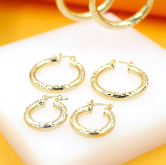 Four polished gold hoop earrings in varying sizes rest on a white display surface against a warm orange backdrop.