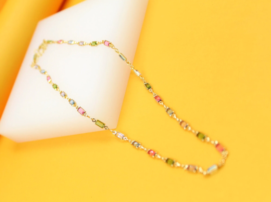 A delicate gold chain bracelet features vibrant, multi-colored baguette crystals resting on a white stand against a bright yellow background.