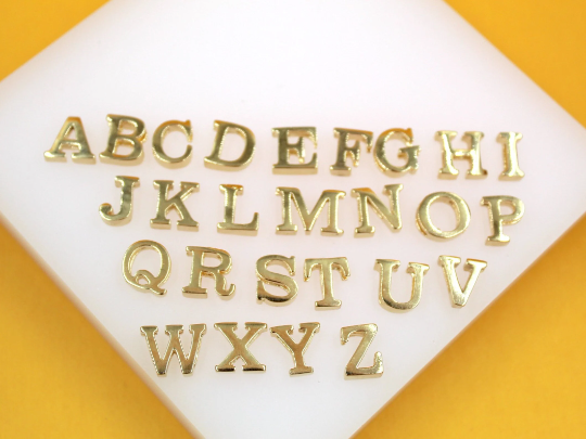 A complete set of shiny gold alphabet letter charms is arranged in alphabetical order on a white display surface against a vibrant orange background.