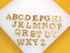 A complete set of shiny gold alphabet letter charms is arranged in alphabetical order on a white display surface against a vibrant orange background.