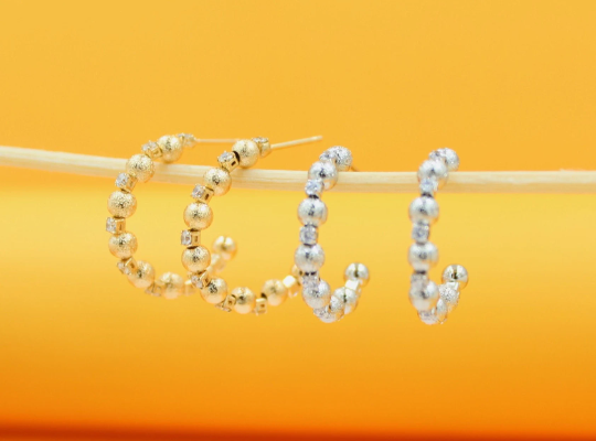 Two pairs of textured gold and silver hoop earrings hang from a wooden stick against a bright yellow background.