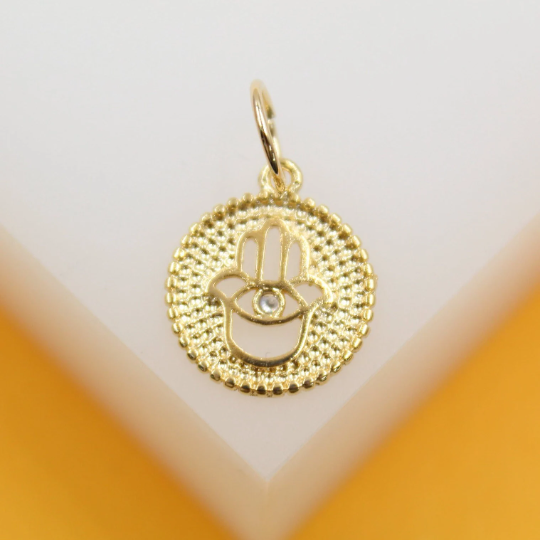 A gold hamsa hand pendant rests on a clean white surface, showcasing its intricate beaded border and central eye detail.