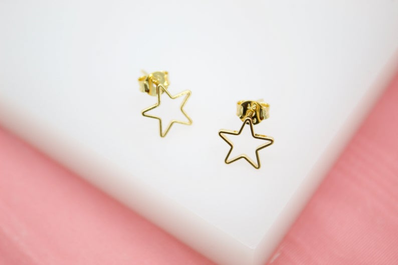 A pair of delicate gold star-shaped stud earrings rests against a white and pink background.