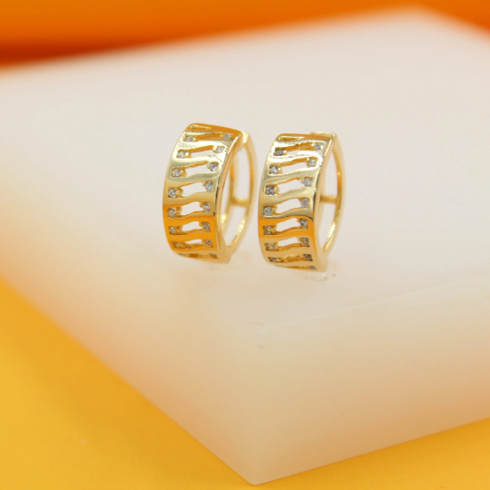 A pair of gold huggie hoop earrings featuring a wavy cut-out pattern encrusted with tiny sparkling diamonds rest on a white display block.
