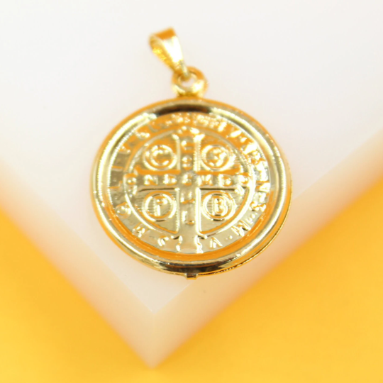 A gleaming gold Saint Benedict medal pendant rests on a white display block against a yellow background.