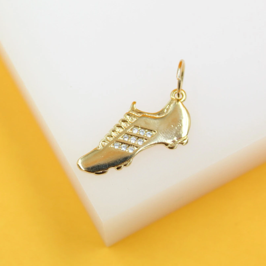 A gold soccer cleat pendant rests on a white display block against a yellow background, showcasing its pave-set cubic zirconia stones along the side stripes.