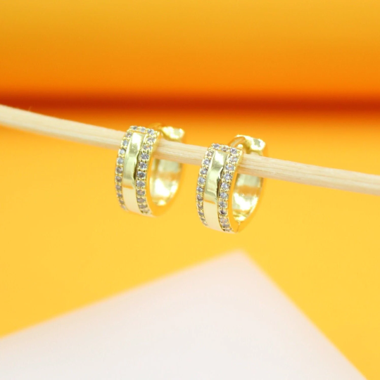 Two gold hoop earrings embellished with tiny sparkling stones hang delicately from a wooden stick against a bright yellow background.
