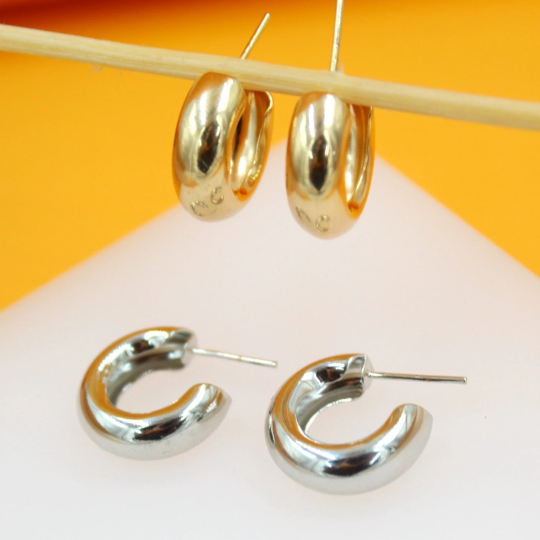 Two pairs of chunky small hoop earrings, one in gold and one in silver, rest on a white surface against an orange background.