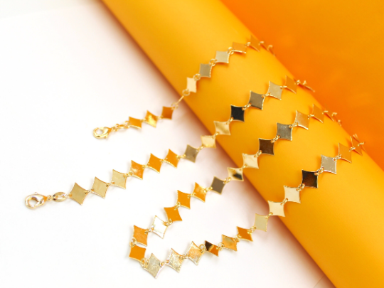 A gleaming gold chain necklace featuring polished diamond-shaped stations drapes gracefully over a vibrant orange display cylinder.