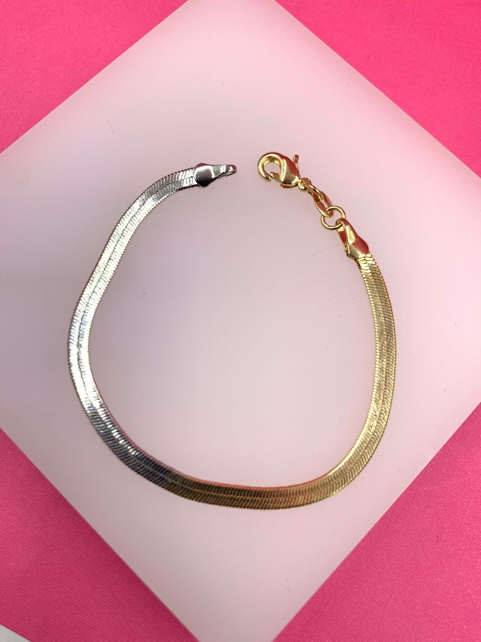A two-tone herringbone chain bracelet made of alternating gold and silver links rests on a white display surface against a vibrant pink background.