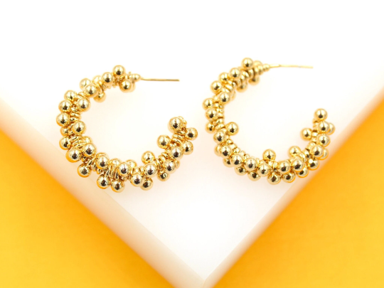 Two textured gold hoop earrings featuring clusters of small spherical beads rest on a white display surface against a vibrant yellow background.