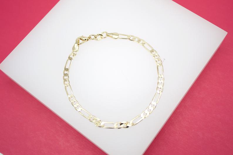 A delicate gold Figaro link chain bracelet rests on a white display surface against a vibrant pink background.