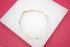 A delicate gold figaro chain bracelet rests on a white display surface against a vibrant pink background.