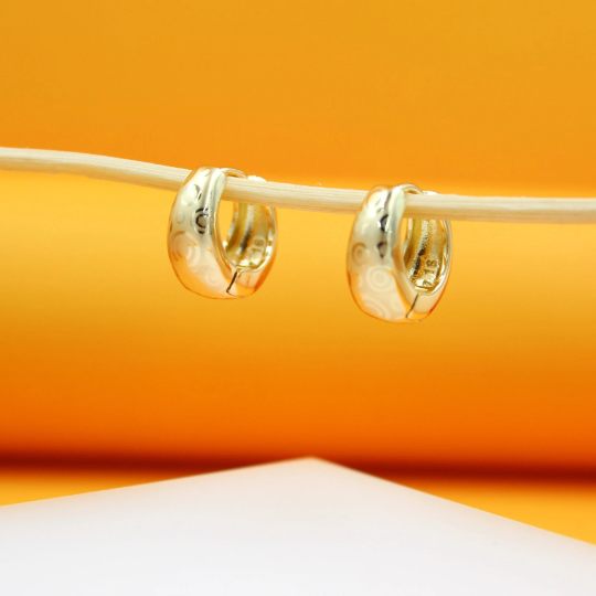 Two engraved gold hoop earrings hang delicately from a thin stick against a warm orange backdrop.