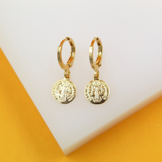 A pair of gold-plated Saint Benedict medallion hoop earrings rest on a white display surface against a vibrant yellow backdrop.