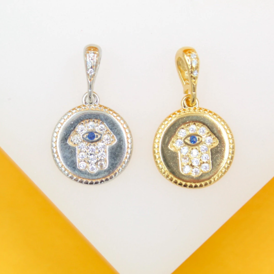 Two circular Hamsa hand pendants with crystal details and blue evil eye centers, one in silver and one in gold, rest side by side on a display stand.