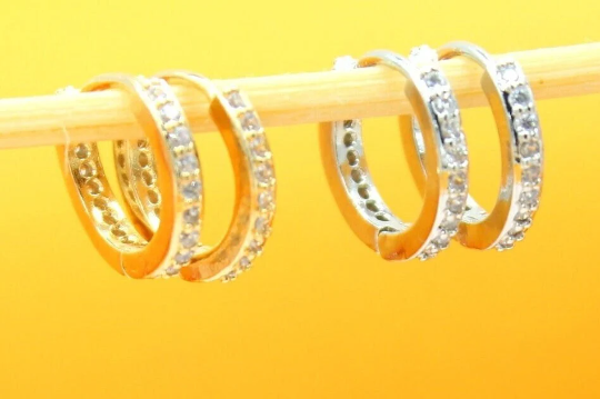 Two pairs of sparkling diamond-encrusted huggie hoop earrings, one in gold and one in silver, hang delicately from a wooden stick against a bright yellow background.