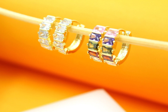 Two pairs of sparkling gold huggie hoop earrings featuring emerald-cut cubic zirconia stones rest on a thin wooden stick against a warm orange background.