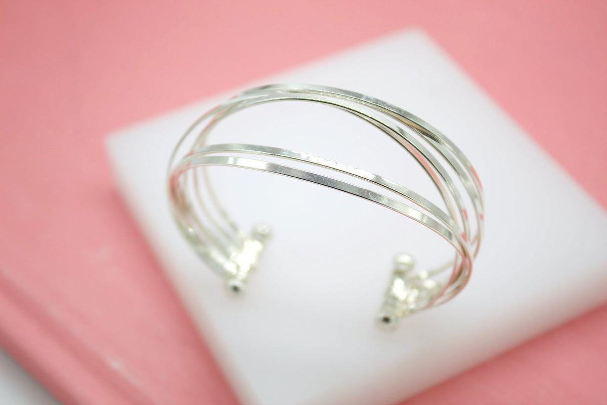 A sleek multi-strand silver cuff bracelet rests on a white display stand against a soft pink background.