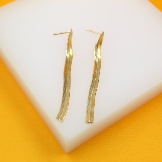 A pair of long, wavy gold herringbone chain earrings rests on a white display stand against a bright yellow background.