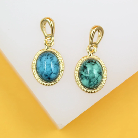 Two gold-framed oval pendants featuring blue and green turquoise gemstones rest side by side against a white and yellow background.