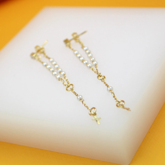 A pair of delicate gold chain earrings features small white pearls and tiny cross charms resting on a white display surface.