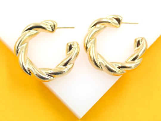 A pair of shiny gold twisted hoop earrings rest on a white surface against a bright yellow background.