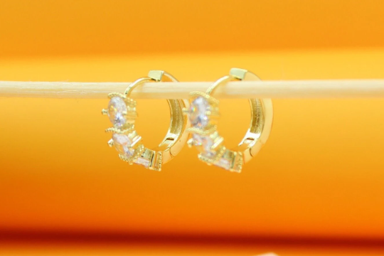 Two gold hoop earrings embedded with sparkling crystals hang delicately from a wooden stick against an orange background.